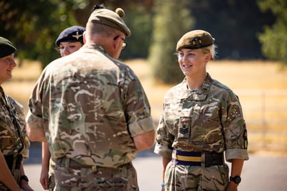 A group of Army Cadets in camouflage uniforms stand outdoors, with one smiling woman facing a man who appears to be speaking to her. Trees and a fence are visible in the background.