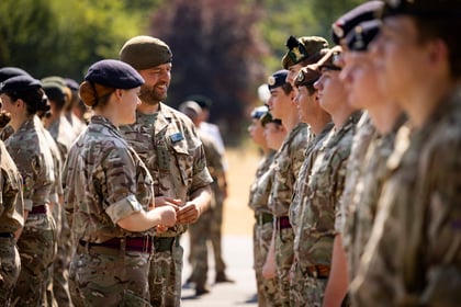 A group of Army Cadets in camouflage uniforms stand in two facing lines outdoors, with two individuals in the foreground smiling and talking to each other. Trees and sunlight are visible in the background.
