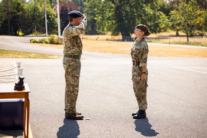 A person in military uniform salutes a cadet outside on a sunny day, with trees and a flag visible in the background.