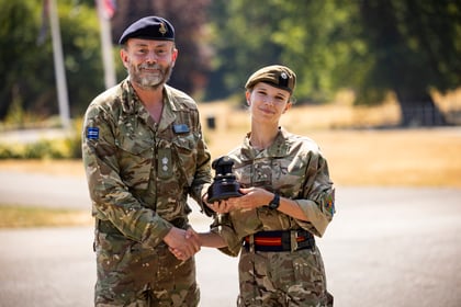 A man and a female cadet in military uniforms shake hands outdoors; the cadet holds a small black trophy. Both are smiling, with trees and flags blurred in the background on a sunny day.