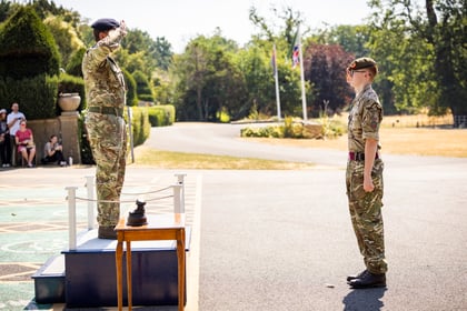 An officer in uniform salutes from a platform whilst a cadet stands to attention outside on a sunny day, with trees and flags in the background.
