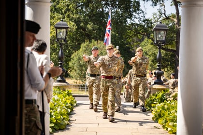 A group of Army Cadets in uniform march in formation on a stone path, with one holding a British flag. People standing nearby applaud. Green hedges and trees are in the background.