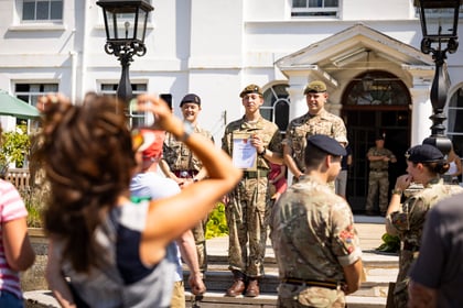 A group of Army Cadets in uniform stand outside a building as people in casual clothes take photos of them on a sunny day. One cadet holds a certificate and all appear to be smiling.