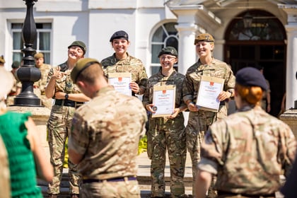 Four Army Cadets in uniform stand smiling and holding certificates outdoors, while others in uniform face them, some taking photos. A white building and a lamp-post are visible in the background.