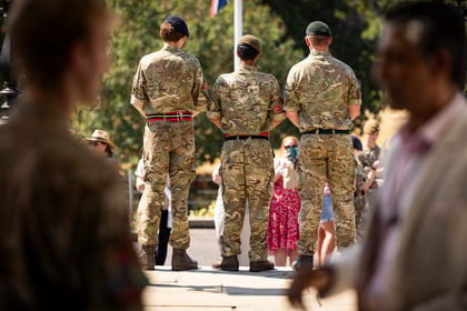 Three Army Cadets in camouflage uniforms stand with their backs to the camera, facing a small group of people outdoors. Some people are blurred in the foreground. The scene appears to be in daylight.