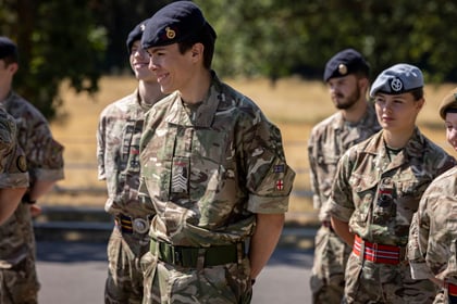 A group of Army Cadets in camouflage military uniforms and berets stand outdoors, some smiling, on a sunny day with trees and dry grass in the background.