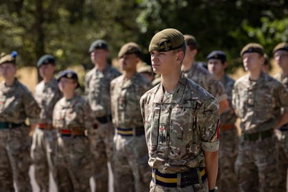 A group of Army Cadets stands outdoors in daylight. One soldier is in focus at the front, whilst others stand blurred in the background. Trees and sunlight are visible.