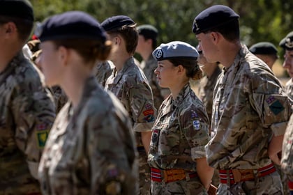 A group of Army Cadets in camouflage uniforms and berets stand outdoors in formation. One woman in a light blue beret stands among them, looking forward. Trees and sunlight are in the background.