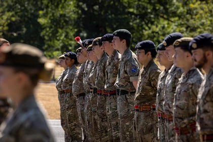 A group of Army Cadets stand in formation outdoors on a sunny day, with trees in the background.