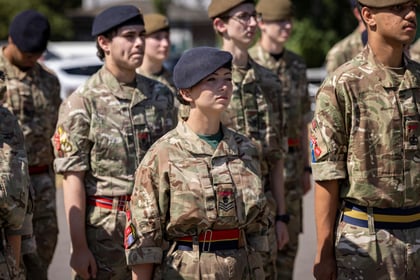 A group of Army Cadets in military uniforms stands in formation outdoors, focusing ahead. Most wear berets and have colourful belts. Trees and blurred buildings are visible in the background.