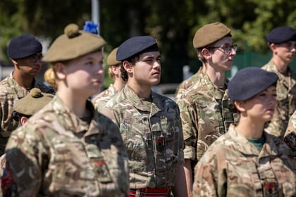 A group of young people in camouflage uniforms and various berets stand outdoors in formation, looking ahead on a sunny day.