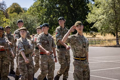 A group of young people in camouflage uniforms and berets march in formation outdoors, with one person at the front saluting. Trees and a fence are visible in the background.