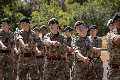 A group of young people in Army Cadet uniforms perform a drill outdoors, extending their arms forward in unison. Trees and greenery are visible in the background.