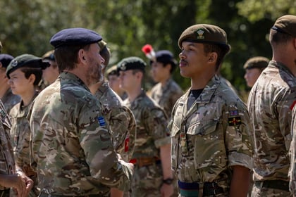 A group of Army Cadets in camouflage uniforms stand outdoors. One man in a beret faces another, engaging in conversation, whilst others stand in rows behind them. Trees are visible in the background.