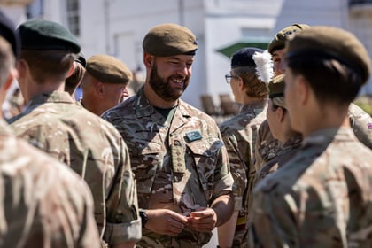 A group of Army Cadets stand together outdoors, talking and smiling. One man in the centre, wearing a beret, is facing others and appears to be the focus of the group. Sunlight brightens the scene.
