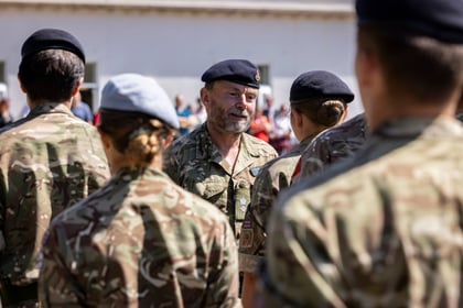 A man in military uniform, wearing a beret, stands facing a group of soldiers outdoors. The other soldiers have their backs to the camera. People and a building are in the background.