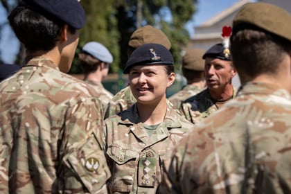 A group of cadets in camouflage uniforms and berets stand outdoors in sunlight, talking and smiling. Trees and a building are visible in the background.