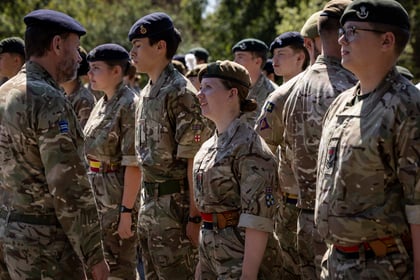 A group of Army Cadets stand in formation outdoors, while an officer in a beret speaks to a female cadet in the front row. Sunlight filters through trees in the background.