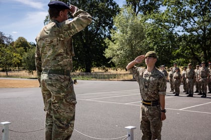 A uniformed officer stands on a platform saluting an Army Cadet who returns the salute; more cadets stand in formation in the background on a sunny day.