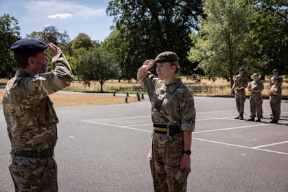A man and woman in military uniforms salute each other outdoors whilst five other uniformed people stand in a line in the background. Trees and dry grass are visible behind them.