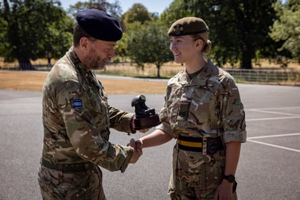 A man in military uniform shakes hands with a smiling woman in Army Cadet uniform, whilst presenting her with a small black trophy outdoors on a sunny day.
