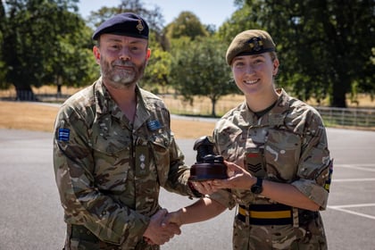 Two people in Army Cadet uniforms shake hands outdoors. The person on the right, smiling, holds a small trophy shaped like a boot. Trees and a field are visible in the background.