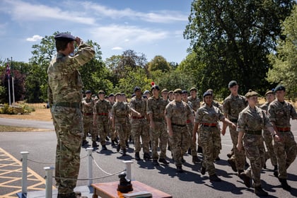 A group of Army Cadets in camouflage uniforms marches in formation outdoors while an officer in uniform salutes them. Trees and a clear sky are in the background.