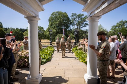 Army Cadets in uniform march under a white colonnade as people on both sides, some in military uniform and others as civilians, applaud. Trees and greenery are visible in the background on a sunny day.