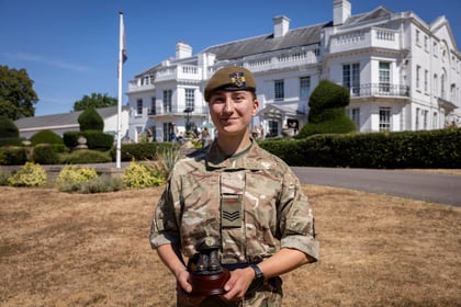 A smiling Army Cadet in camouflage uniform stands holding a trophy in front of a large white building with manicured gardens and a flagpole on a sunny day.
