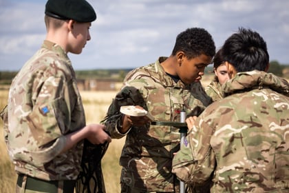 Four young people in camouflage uniforms work together outdoors, assembling equipment in a grassy field under a partly cloudy sky. One holds a small dish whilst others focus on the task.
