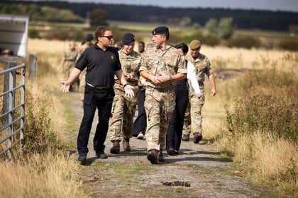 A group of people, some in uniforms and others in civilian clothing, walk and talk along a rural path surrounded by grass and fields on a sunny day.