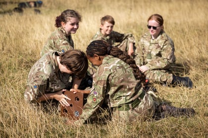 Five young people in camouflage uniforms sit on dry grass, focused on a wooden puzzle box. Two are actively working on the puzzle, while the others watch and smile in the background.