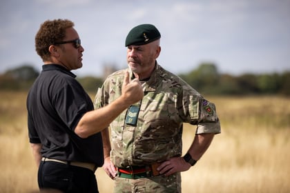 A man in black gestures while speaking to a man in camouflage military uniform and green beret, standing outdoors in a grassy field under a cloudy sky.
