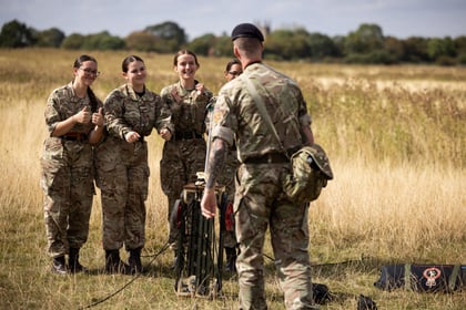Four people in Army Cadet uniforms smile and give thumbs up whilst standing in a grassy field as another uniformed person with a rucksack faces them. Equipment is set up nearby and trees are visible in the background.