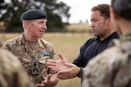 Two men, one in military uniform and the other in a black polo shirt, have an animated conversation outdoors whilst soldiers stand nearby, blurred in the foreground.