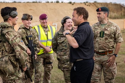A group of Army Cadets in camouflage talk and laugh outdoors with a man in black. One person wears a yellow vest, another wears a maroon beret, and everyone appears relaxed and happy.