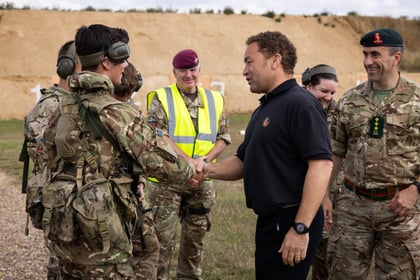 A man in a black shirt shakes hands with a cadet in camouflage, surrounded by other personnel outdoors. They all appear to be smiling and engaged in friendly conversation.