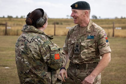Two senior personnel in camouflage uniforms talk and smile outdoors on grassy terrain. One wears ear protection, while the other, adorned with a beret and various badges, stands nearby. A fence and field are visible in the background.