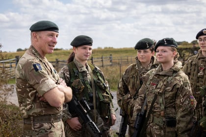 A senior military officer in uniform talks with four young Army Cadets in camouflage and berets outdoors. The group stands on a grassy field under a partly cloudy sky, holding rifles and listening attentively.