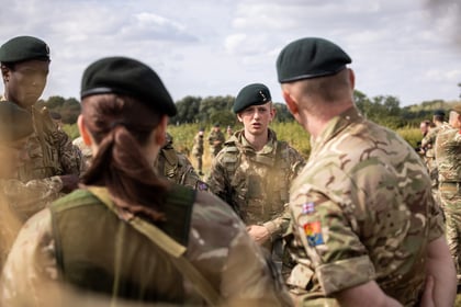 A group of Army Cadets in camouflage uniforms and green berets stand outdoors, gathered in conversation. The background shows more cadets and greenery under a partly cloudy sky.