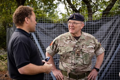 A man in military camouflage with a beret speaks with another man in a black shirt outdoors, standing in front of a wire fence and trees.