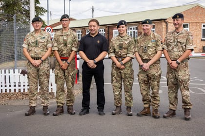Six people, five in Army Cadet uniforms and one in black clothing, stand side by side outside on a paved area near a brick building and a white picket fence, smiling at the camera.