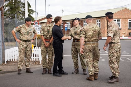 Six people, five in uniforms and one in black clothing, stand outside talking near a brick building. Two of them are shaking hands. There are road signs and a white picket fence in the background.