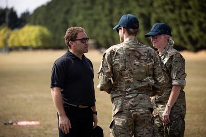 A man in a black shirt talks to two people in camouflage uniforms outdoors on a grassy area, with trees in the background.
