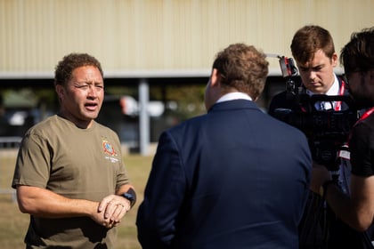 A man in a brown shirt speaks outdoors to a man in a suit as a camera crew films them, with a beige building and grass visible in the background.