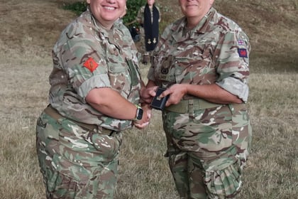 Two women in military uniforms shake hands outdoors during a ceremony, both smiling. One woman holds a small black case. Trees and grass are visible in the background, with another person standing further back.