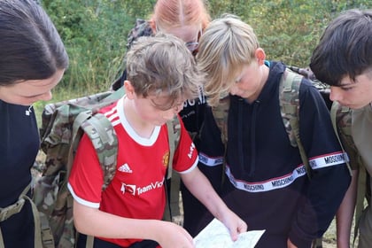 Five teenagers with rucksacks gather outdoors, closely examining a paper map together. They appear focused and engaged, surrounded by greenery and casual outdoor clothing.