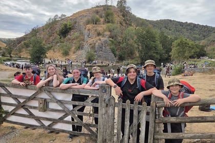 A group of smiling children with rucksacks and hats stand by a wooden gate, with a crowd, rocky hill, and trees in the background under a cloudy sky.