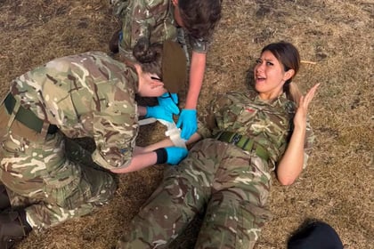 Two people in camouflage uniforms and blue gloves bandage the knee of a woman in uniform lying on dry grass, looking surprised. A black beret lies nearby. Trees and a road are in the background.
