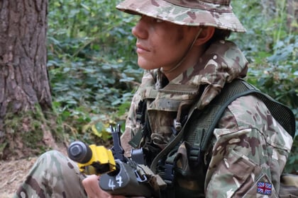 A person in camouflage military uniform and hat sits in a forest, holding a black and yellow training rifle. The uniform has British flag and patch insignia on the arm.